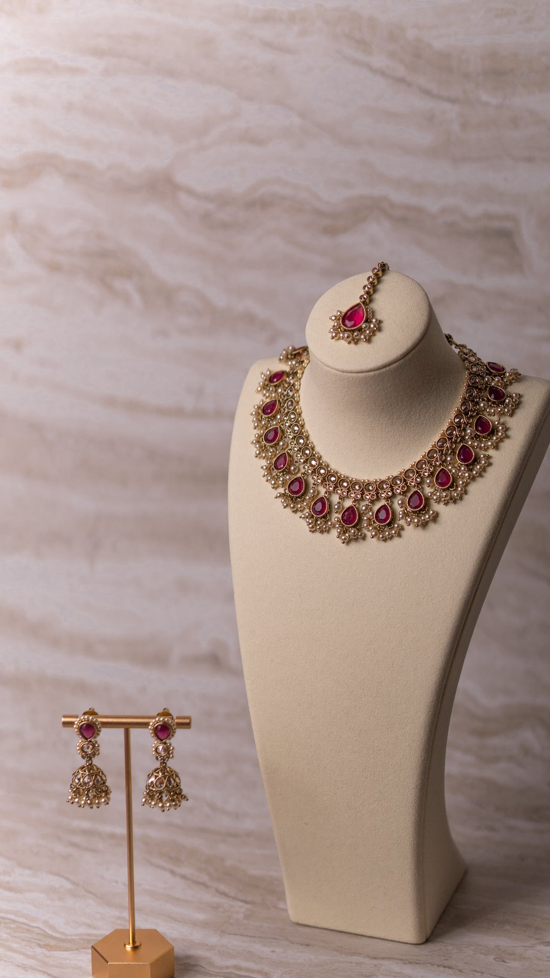 Gold necklace with red stones on a mannequin bust and matching earrings on a stand against a marble background.