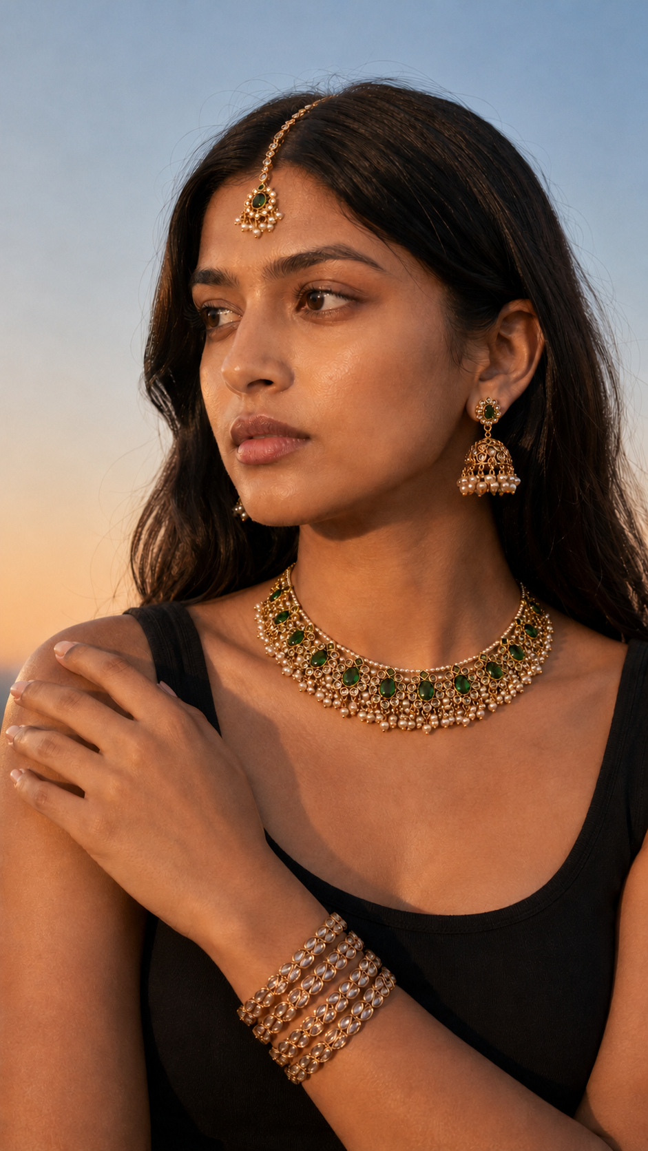 A woman wearing a gold-toned traditional jewelry set with green stone accents, including a necklace, earrings, and stacked bangles, paired with a black sleeveless top against a soft sky background.